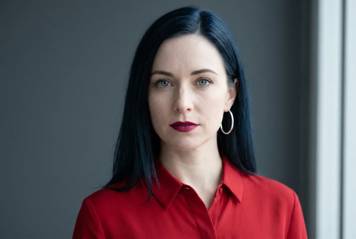 Woman with blue-black hair, porcelain skin, and icy blue eyes in a true-red blouse and silver hoop, on charcoal-grey.